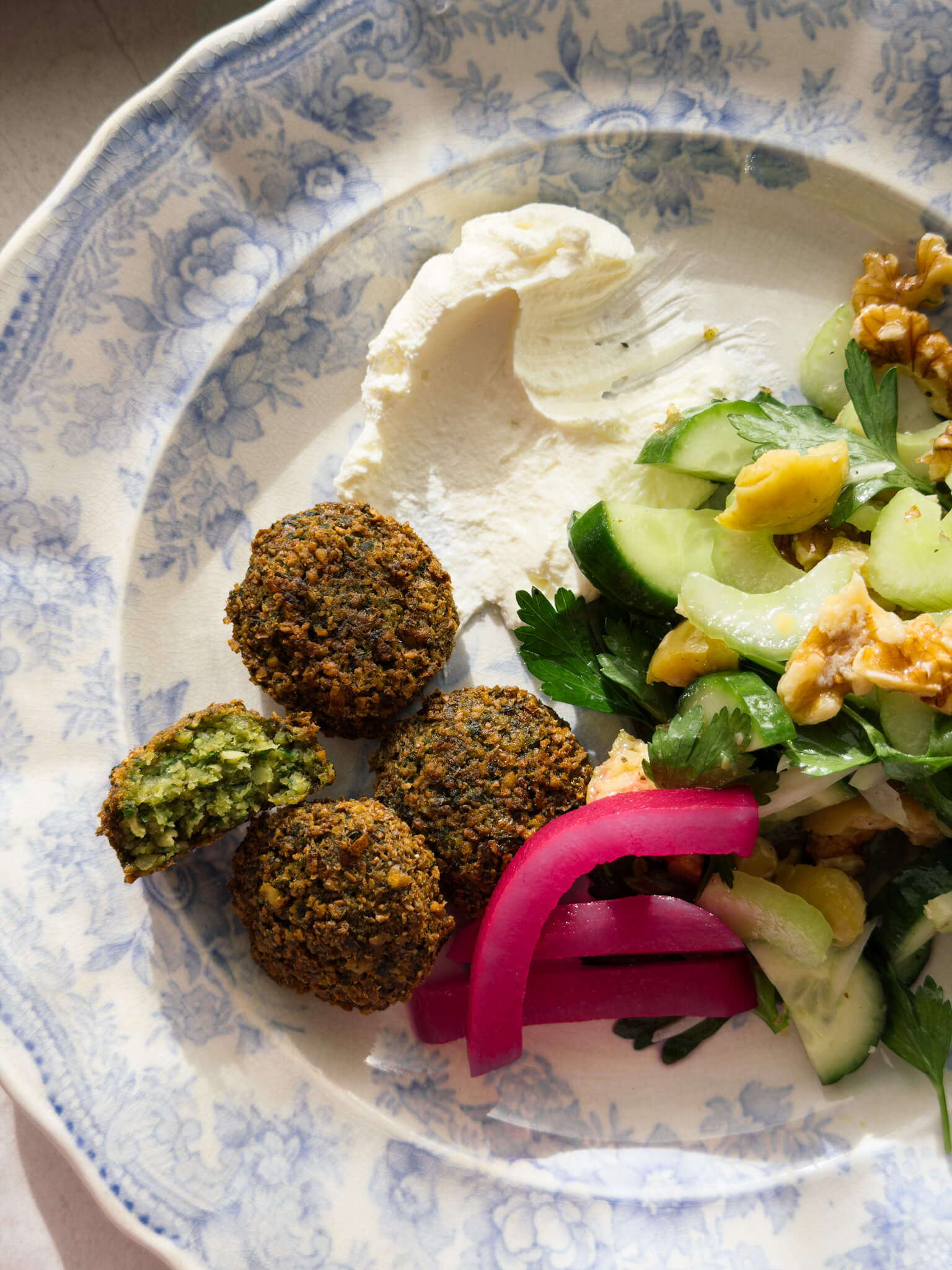 A close up shot of a blue patterned plate shot in streaming sunlight. On the plate are four freshly cooked falafels with the mid-section of one viewable, revealing a herby green centre. On the plate is fresh labneh, pink pickled turnips, and a crisp green salad. 