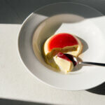 A white dessert bowl with a layered blood orange jelly and panna cotta pictured in direct sunlight. A spoonful of the dessert is set onto the plate showing the distinct layers.