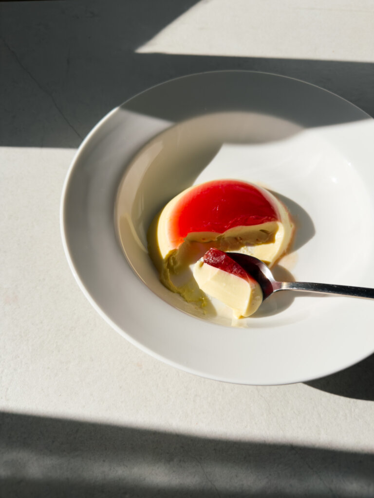 A white dessert bowl with a layered blood orange jelly and panna cotta pictured in direct sunlight. A spoonful of the dessert is set onto the plate showing the distinct layers.