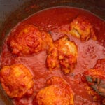 A close up photo of chicken thighs simmering in a tomato sauce, with bay leaves, all spice and cinnamon.
