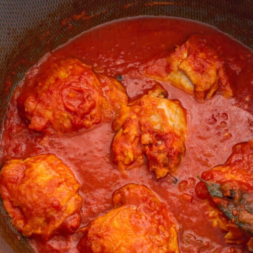 A close up photo of chicken thighs simmering in a tomato sauce, with bay leaves, all spice and cinnamon.