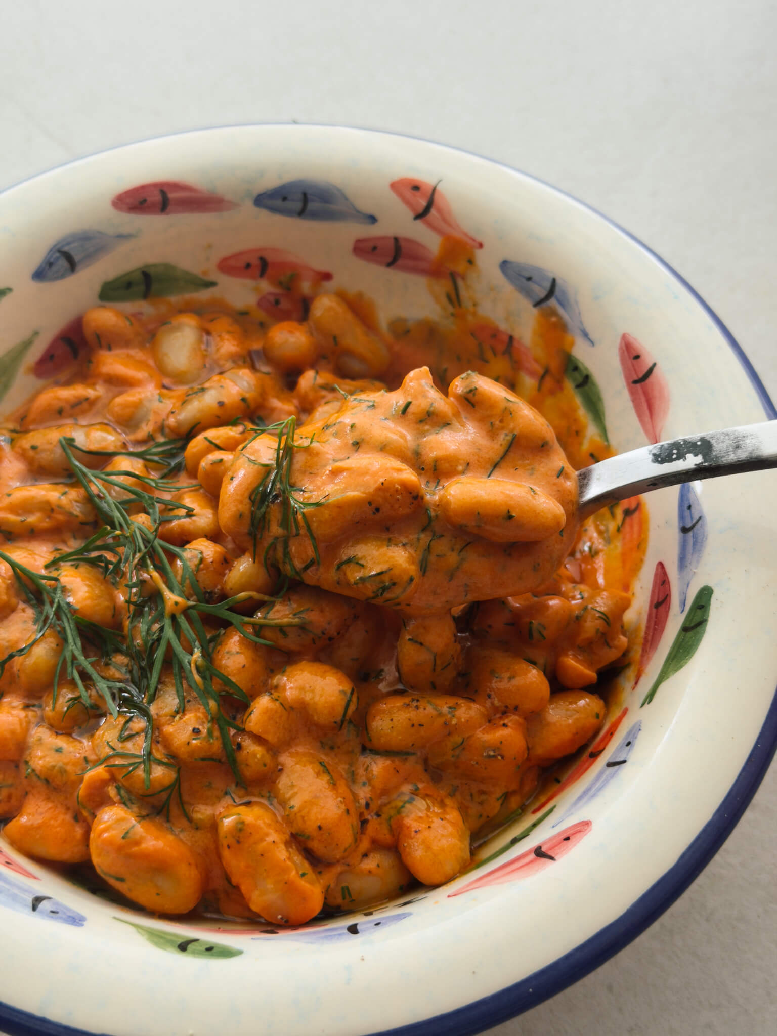 A close up photo of a decorative bowl with hand painted colourful fish and a blue rim, containing creamy tomato beans topped with olive oil and fresh dill and a spoon lifting some of the beans out of the bowl.