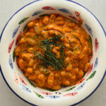 A top down photo of a decorative bowl with hand painted colourful fish and a blue rim, containing creamy tomato beans topped with olive oil and fresh dill.