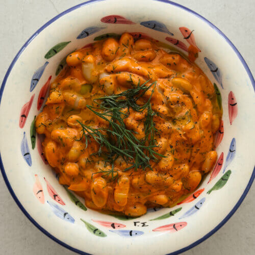 A top down photo of a decorative bowl with hand painted colourful fish and a blue rim, containing creamy tomato beans topped with olive oil and fresh dill.