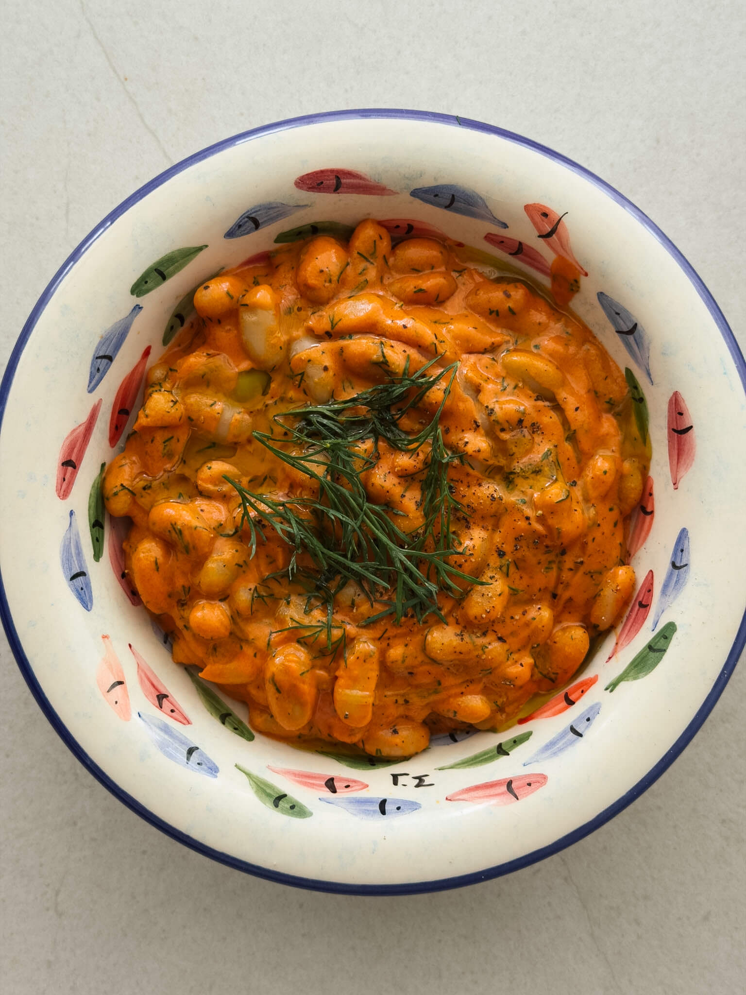 A top down photo of a decorative bowl with hand painted colourful fish and a blue rim, containing creamy tomato beans topped with olive oil and fresh dill.