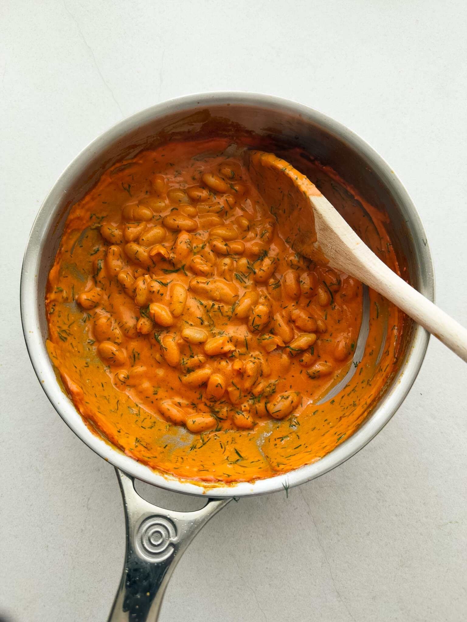 a top down photo of a stainless steel sauté pan containing a vibrant orange, creamy tomato cannellini bean dish, spiked with fresh, finely chopped dill.